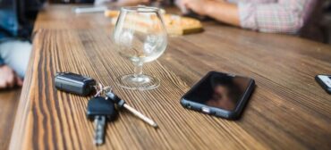 Kluczyki samochodowe, pusty kieliszek i smartfon leżące na drewnianym stole, z rozmytymi osobami siedzącymi w tle.